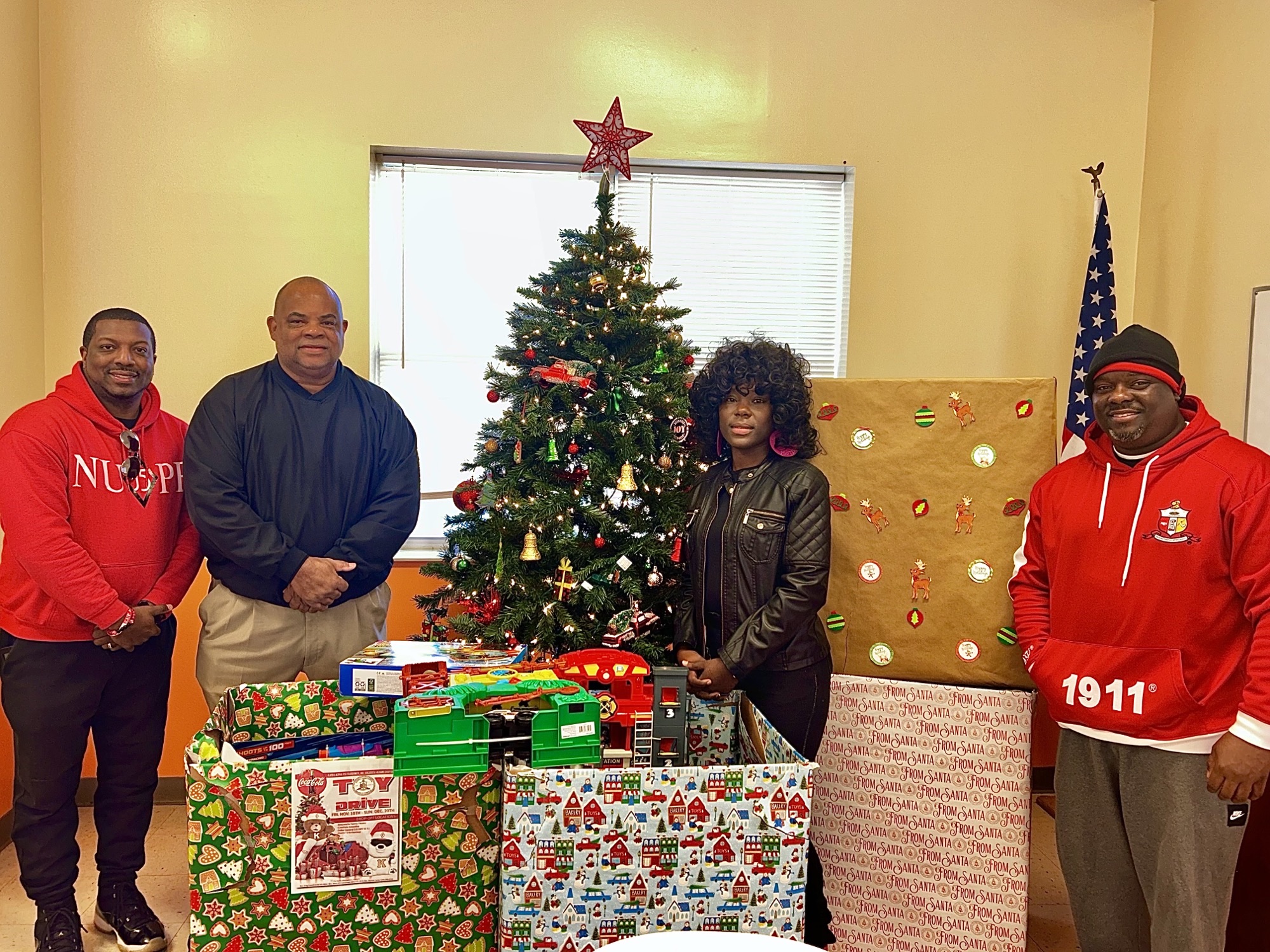 several individuals posing near toy drive boxes and Christmas tree